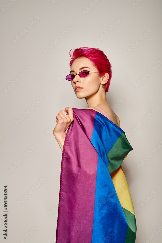 confident young woman with pink hair and sunglasses posing with lgbt rainbow flag on grey backdrop