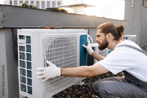 Adult bearded construction worker crouching and keeping hands on air conditioner while checking serviceability. Caucasian qualified man wearing gloves examining device on roof of factory. photo