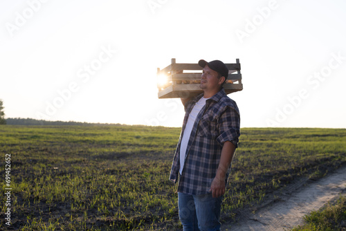 A tired worker carries a box of potatoes on his shoulder along the road along the field in the rays of the setting sun A man works in the field and harvests potato vegetables. End of the working day.