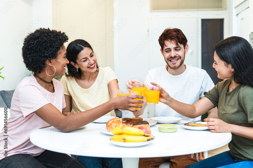 Four multi-ethnic friends laugh and toast at a home breakfast with fresh juice and fruit.