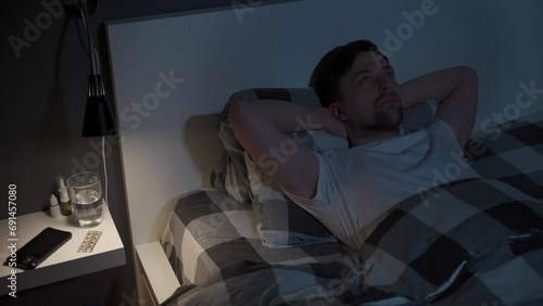 Young man looks at the ceiling at night while lying in bed instead of sleeping, checking the time on his mobile phone, suffering from insomnia. Displeased male lying in bed unable to fall asleep. photo