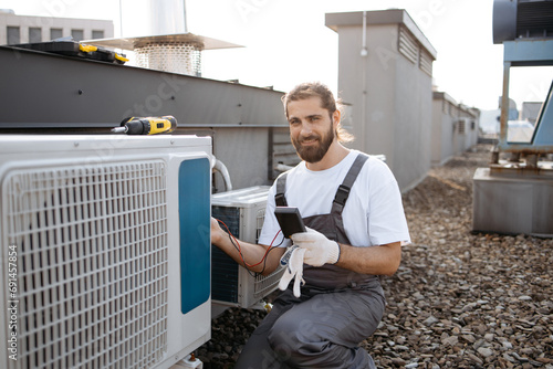 Concentrated man squatting down and holding voltage tester while applying to air conditioner. Builder man closed in overalls performing work and checking serviceability of cooling system.