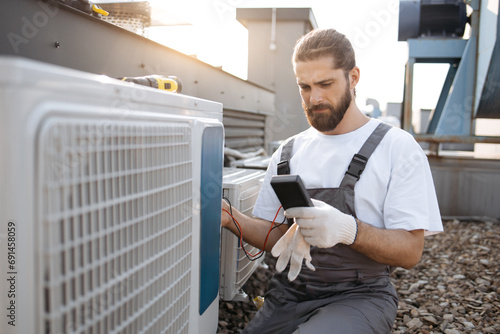 Focused bearded master wearing gloves holding digital multimeter and checking voltage level in air conditioner. Caucasian man working with factory equipment on open air terrace. photo