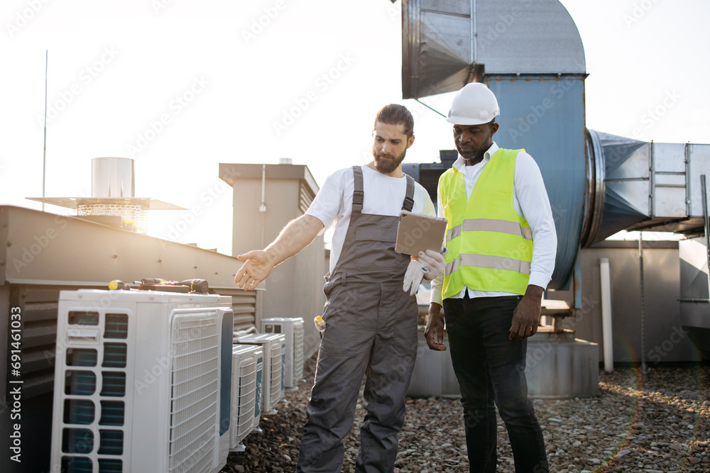 African american supervisor and caucasian technician standing next to each other and looking at modern tablet screen. Qualified factory workers reading information about air conditioners on rooftop. African american supervisor and caucasian technician standing next to each other and looking at modern tablet screen. Qualified factory workers reading information about air conditioners on rooftop.