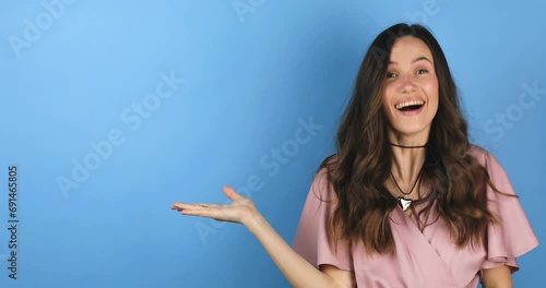 Excited surprised woman look at side, say wow and pointing hands aside isolated on blue background. Brunette girl in pink dress. Look over here. Sale here. Thumb up.