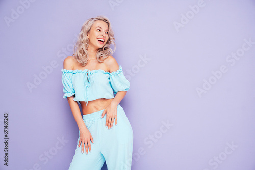 Young beautiful smiling female in trendy summer blue costume clothes. Carefree blond woman posing near violet wall in studio in jacket and shorts. Positive model having fun. Cheerful and happy