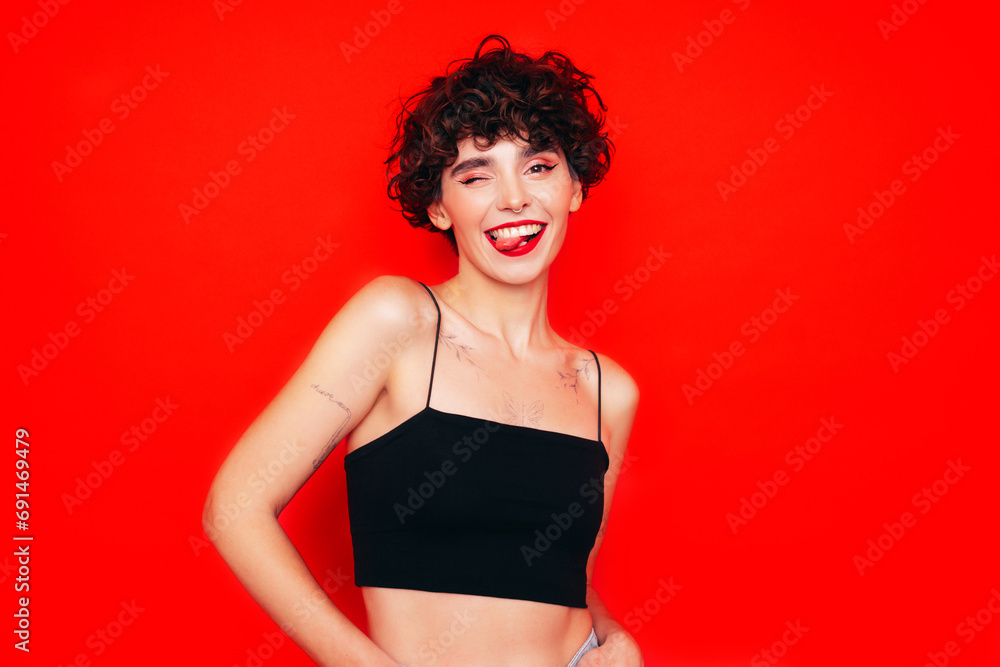 Young beautiful smiling female in trendy summer black tank top. Carefree woman posing near red wall in studio with curly hairstyle. Positive model having fun, going crazy. shows tongue Young beautiful smiling female in trendy summer black tank top. Carefree woman posing near red wall in studio with curly hairstyle. Positive model having fun, going crazy. shows tongue