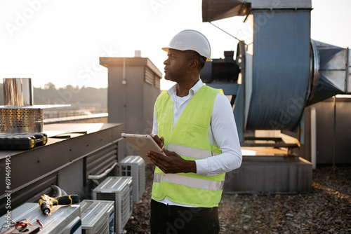 Competent craftsman wearing white hard hat standing and holding wireless tablet in hands outdoors. Black concentrated man searching for information in device and monitoring work at plant in roof.