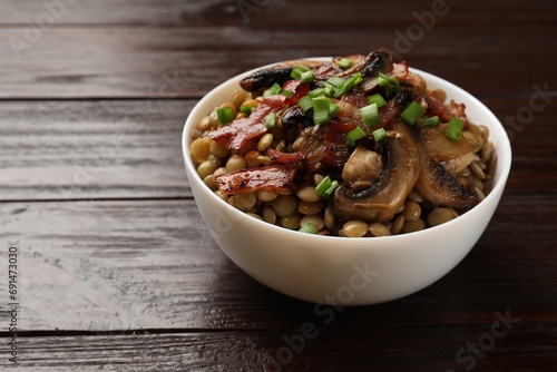 Delicious lentils with mushrooms, bacon and green onion in bowl on wooden table, closeup. Space for text