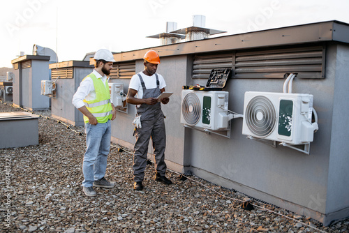 African american technician with tablet in hands showing fixed air conditioner to caucasian manager in white hard hat on rooftop. Factory workers cooperating for maintaining modern equipment. photo