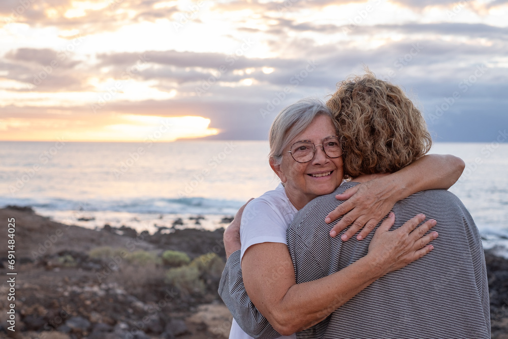 Fototapeta premium Romantic senior couple of women embrace each other on the seashore at sunset light, two embraced people stay together expressing love and tenderness