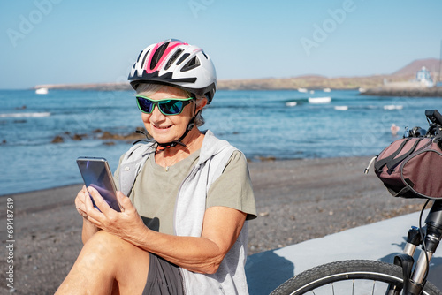 Smiling senior cyclist woman sitting at the beach with electric bike using mobile phone. Authentic retirement living and healthy lifestyle concept.