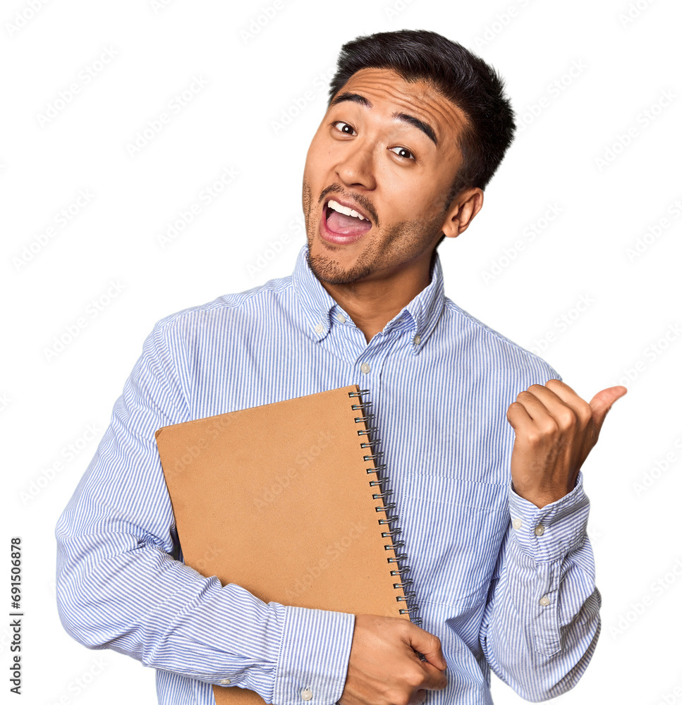 Chinese student with a notebook in studio points with thumb finger away, laughing and carefree. Chinese student with a notebook in studio points with thumb finger away, laughing and carefree.