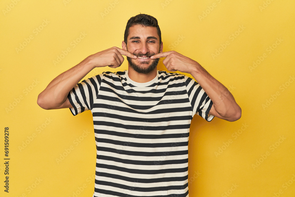 Young Hispanic man on yellow background smiles, pointing fingers at mouth. Young Hispanic man on yellow background smiles, pointing fingers at mouth.