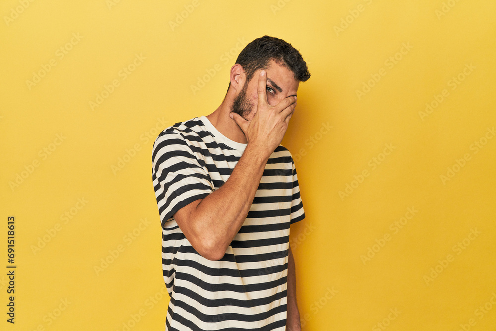 Young Hispanic man on yellow background blink at the camera through fingers, embarrassed covering face. Young Hispanic man on yellow background blink at the camera through fingers, embarrassed covering face.