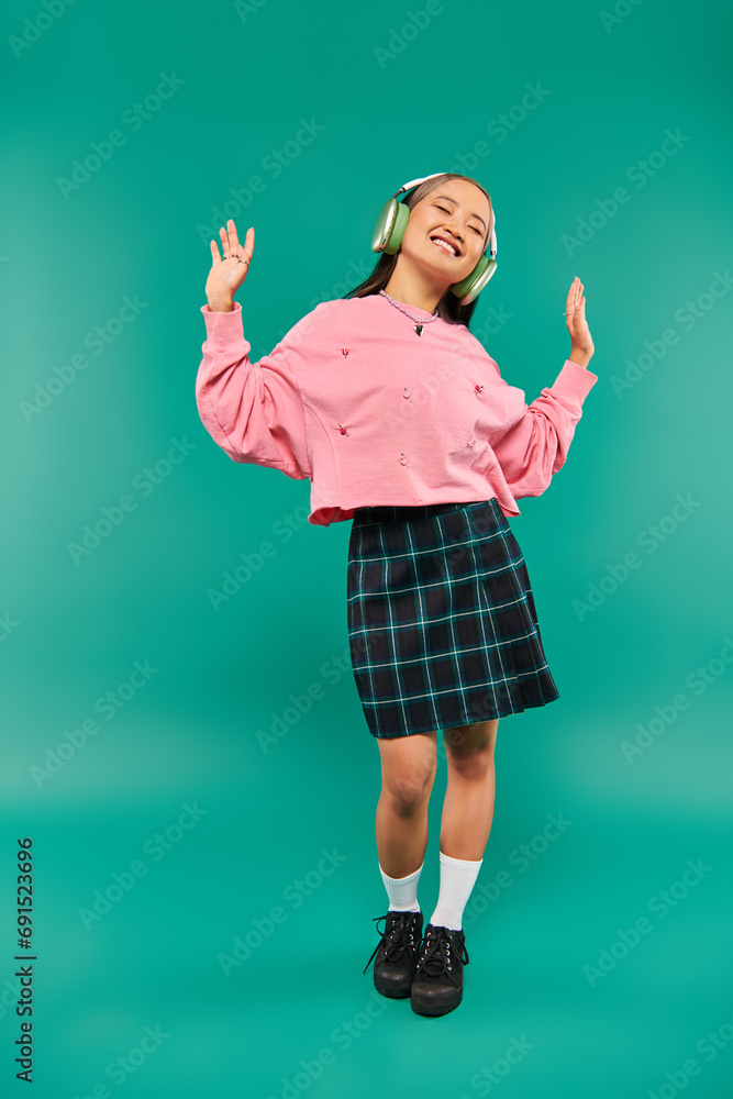 full length of positive asian girl in wireless headphones dancing on turquoise background full length of positive asian girl in wireless headphones dancing on turquoise background