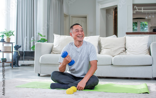 Asian aging senior old man wearing casual comfortable t shirt, sitting on floor in living room at cozy home, doing exercise with happiness, smiling. Healthcare, Retirement Concept.