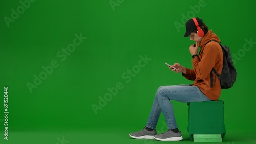 Portrait of person tourist isolated on chroma key green screen background. Young man sitting holding smartphone listening music in headphones.