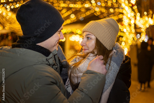 Positive, cheerful couple hugging during x-mas evening. Decorated lights on the streets outdoors (ID: 691532675)