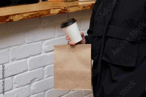 A man in black holds a paper bag and a paper glass of coffee against the background of a bar counter.