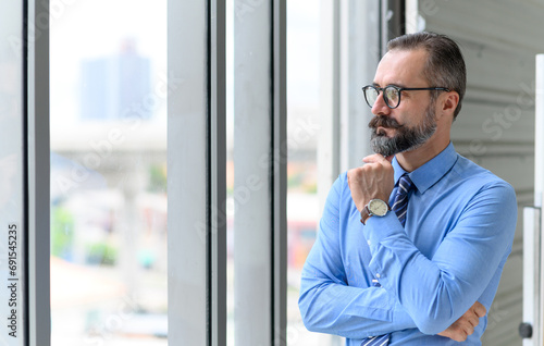 Portrait of confident middle-aged businessman in blue shirt and wearing glasses with a beard and mustache standing next to window in office with copy space.