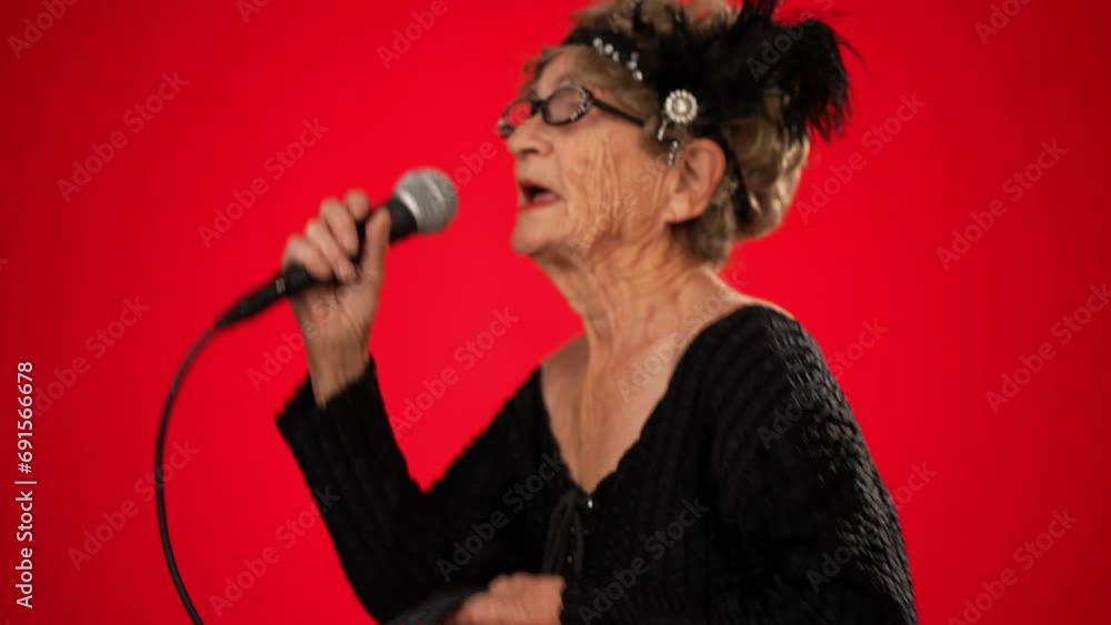 Funny toothless elderly woman in fashionable look with glasses, walks into frame singing enthusiastically into a microphone and dancing isolated on solid red background Funny toothless elderly woman in fashionable look with glasses, walks into frame singing enthusiastically into a microphone and dancing isolated on solid red background