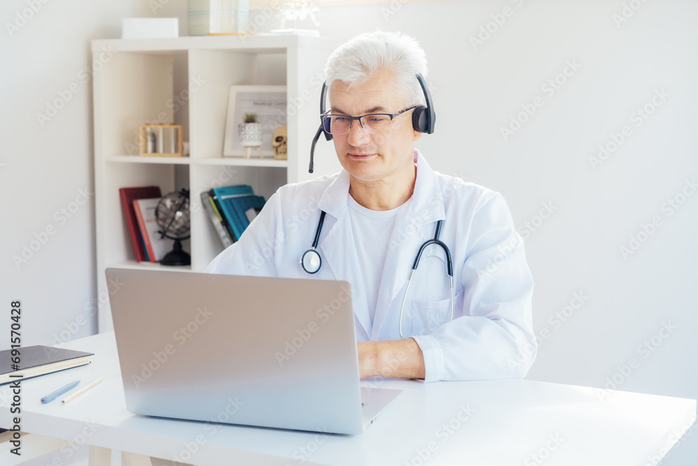 Middle aged male doctor using headset and laptop for online video call consulting of patient. Telemedicine concept for domestic health treatment. Online remote medical appointment. Medical technology Middle aged male doctor using headset and laptop for online video call consulting of patient. Telemedicine concept for domestic health treatment. Online remote medical appointment. Medical technology