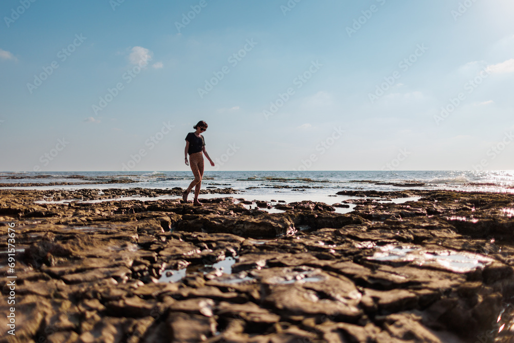 A teenage girl walks alone along the water line on a sand and stone beach during summer vacation. \