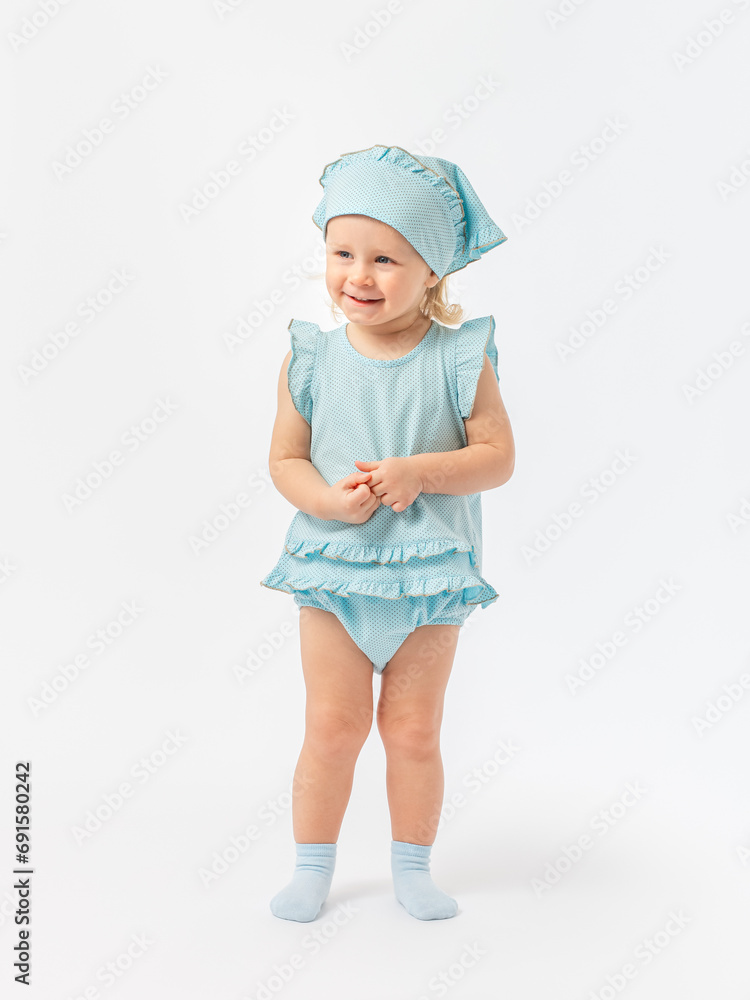 A blonde girl of 2 years old is smiling, touching her fingers and looking away in full height in a blue dress and socks on a white background. A blonde girl of 2 years old is smiling, touching her fingers and looking away in full height in a blue dress and socks on a white background.