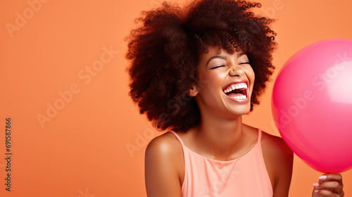 Joyful woman with an afro hairstyle, laughing and holding a pink balloon, wearing a light pink dress against a peach-colored background. © MP Studio