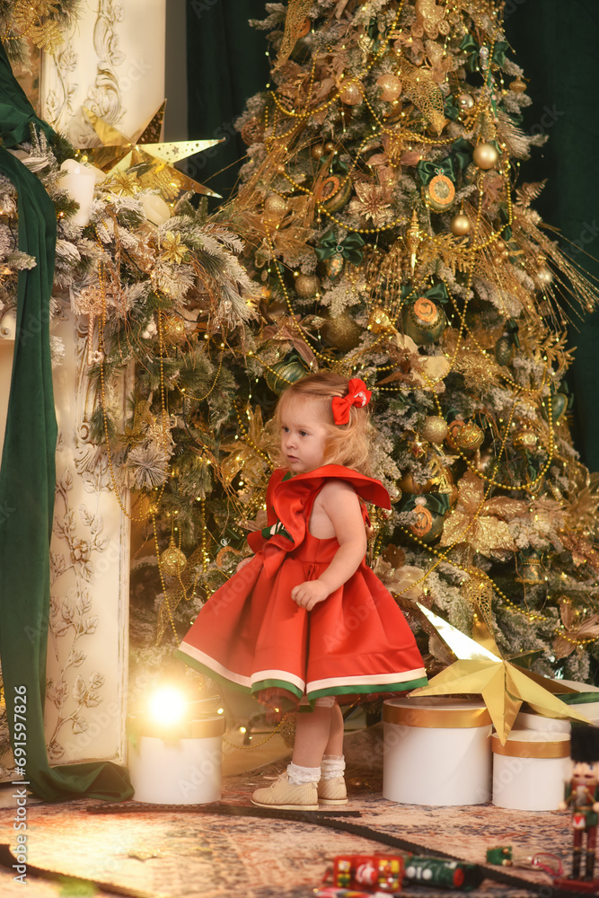 A little beautiful girl plays near the Christmas tree. Beautiful little child in a beautiful red dress