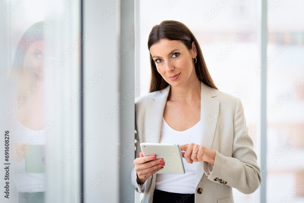 Smiling mid aged businesswoman standing by the window in a modern office and holding touchpad in her hand Smiling mid aged businesswoman standing by the window in a modern office and holding touchpad in her hand
