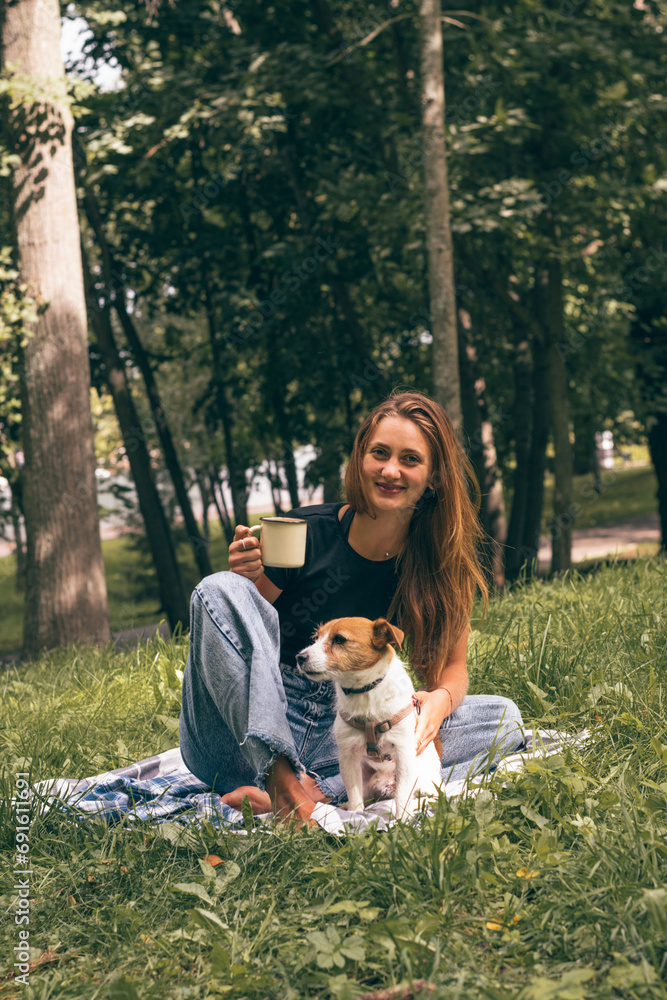 A young positive woman walks with her dog in the park.A woman drinks tea from a thermos.A Jack Russell Terrier dog is sitting next to her.