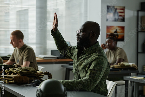 African American guy in military uniform and eyeglasses raising hand and looking at teacher after lecture or presentation to ask question photo