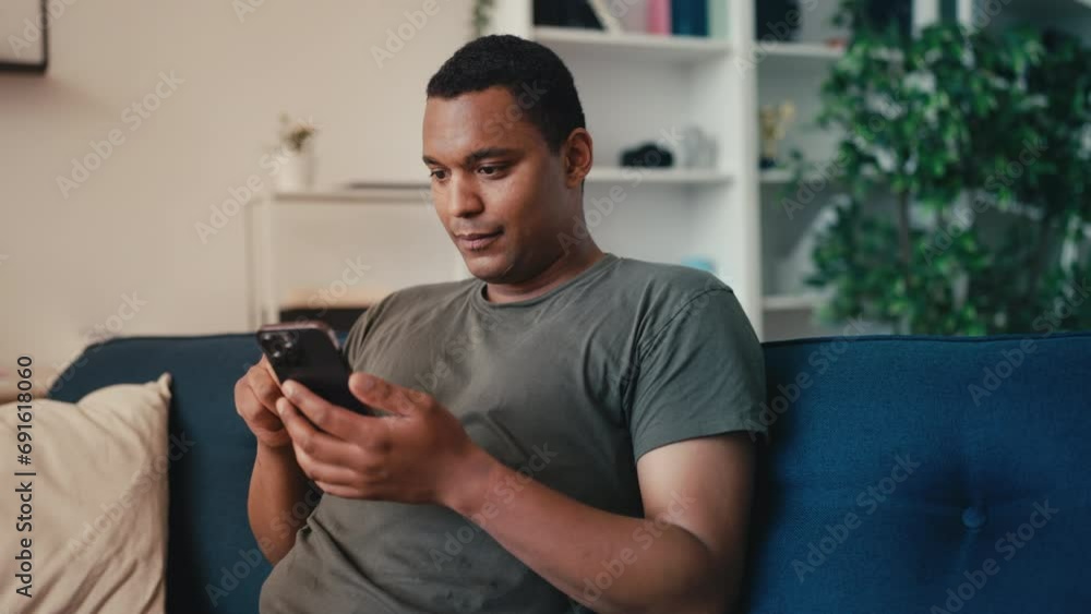 Smiling African American man scrolling phone while sitting on couch at home Smiling African American man scrolling phone while sitting on couch at home