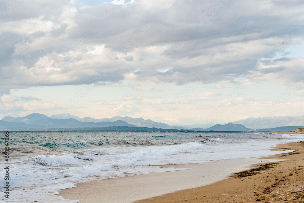 Expansive beachfront with waves and a mountainous backdrop under a cloudy sky