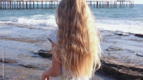 Back view of a blonde little girl eating ice-cream and talking to camera while standing on seashore, concept of child's happiness and freedom