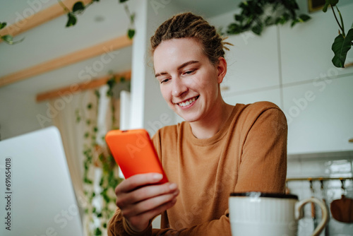 Smiling young woman using smartphone at kitchen table with laptop in daylight photo