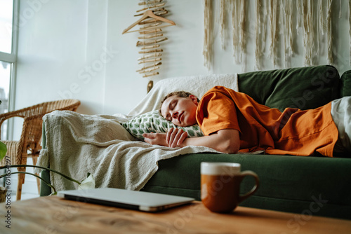 Young woman sleeping on comfortable sofa with cushion at home near laptop and mug photo
