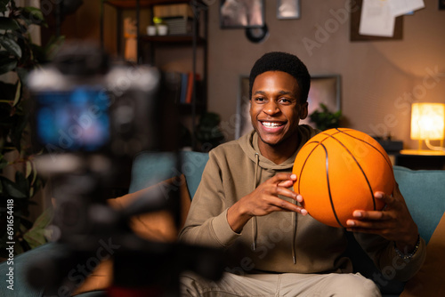 A basketball player sits in a tidy living room, ball in hand, dressed casually. He looks relaxed and focused, taking a break from the game.
