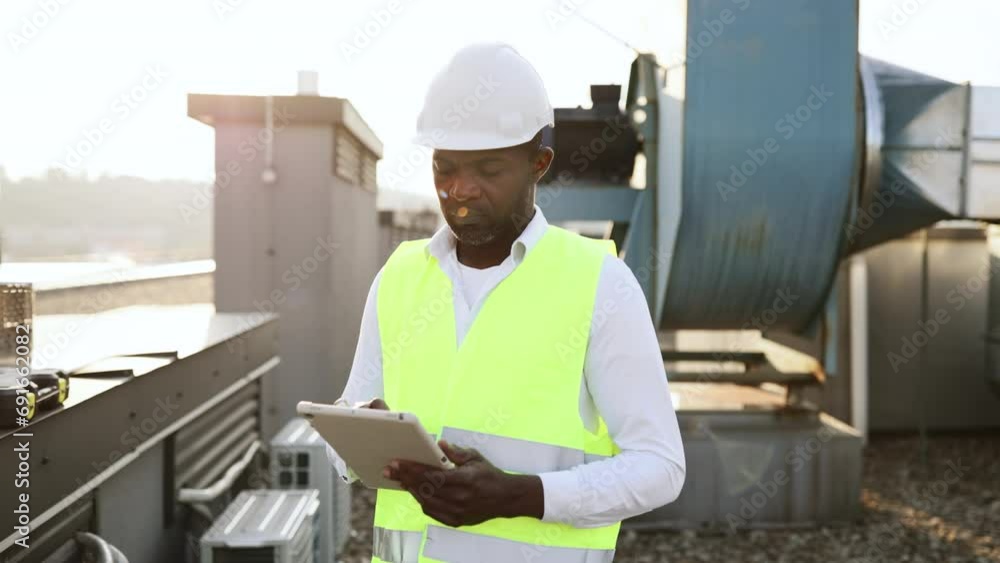 Serious factory worker standing and holding modern tablet in hands while servicing devices at factory. African american man wearing uniform inspecting area while performing work in local roof. Serious factory worker standing and holding modern tablet in hands while servicing devices at factory. African american man wearing uniform inspecting area while performing work in local roof.