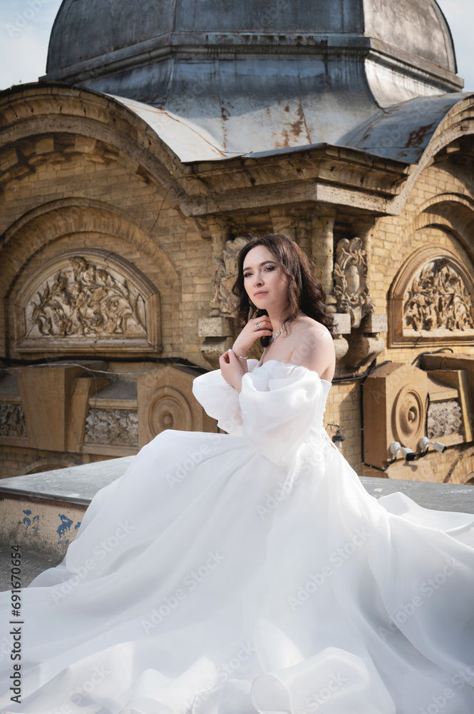 bride in a white dress sits on a ledge with an old building in the background. Young Caucasian woman straightens her hair at a wedding in an ancient city