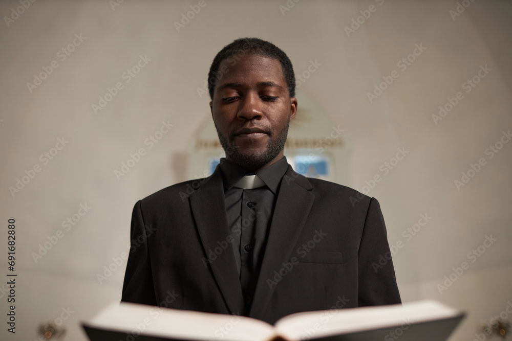 Waist up portrait of young Black priest reading Bible standing at altar during Sunday service Waist up portrait of young Black priest reading Bible standing at altar during Sunday service