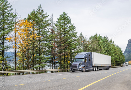 High cab long hauler gray big rig semi truck transporting cargo in dry van semi trailer driving on the wide highway road along the Columbia River with autumn trees on the side