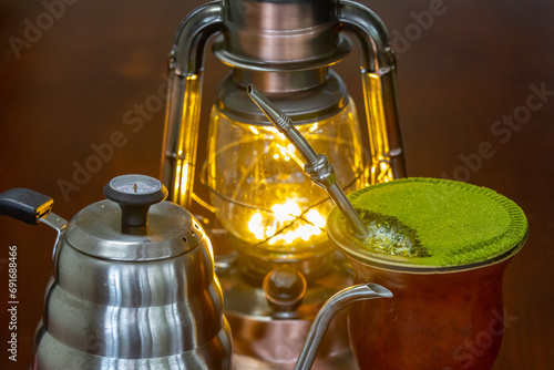 Traditional chimarrão prepared with yerba mate (Ilex paraguariesis) in a porongo gourd, next to a vintage lamp and barista-style gooseneck kettle photo