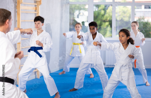 Motivated sporty black girl with group of tweens wearing white kimonos diligently performing kata routines to hone karate techniques in training room..