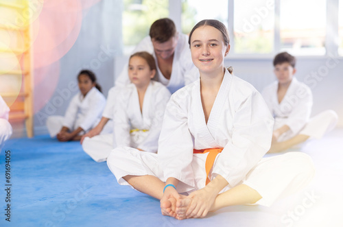 Positive preteen girl in kimono sitting on tatami in gym, performing butterfly stretch during group martial arts training..