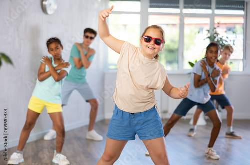 Dynamic little girl training Break-dance poses in dancehall with other attendees of dancing courses