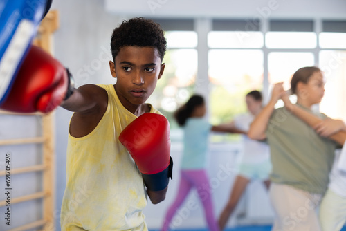 Motivated focused african american boy wearing boxing gloves working out with coach during group self-defense course, practicing punches on mitts.. © JackF