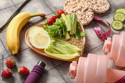 Plate with fresh healthy products and dumbbells on grey tile background, closeup. Diet concept